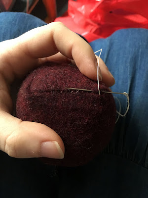 A red wool pincushion, shown from the side, with a needle and white thread inserted into the side seam