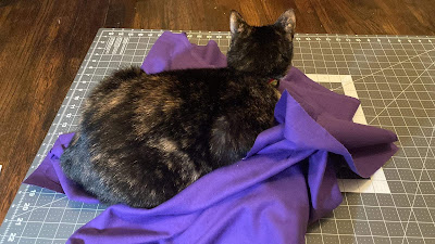 A black and orange tortoiseshell cat is sitting in loaf pose on purple flannel atop a fabric cutting mat, she is facing away from the camera.