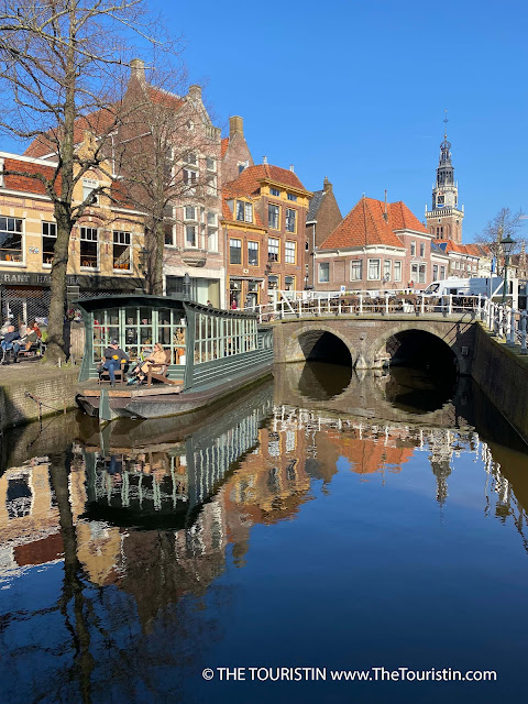 A couple dressed in winter coats and gloves sitting on the porch in the bow of a large flat boat furnished with large green wooden window frames. All this and surrounding red brick houses, a tall church spire and a red brick bridge with its white railing reflect in the quiet canal under a bright blue sky.