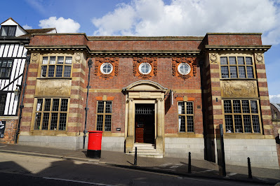 A red brick building with terracotta and light-brown stone details including reliefs around round first-florr windows, stone reliefs under leaded rectangular first-floor windows, and a semi-circular stone pediment above the front door.