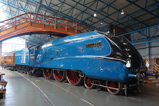 Mallard on the famous trains on display at the National Railway Musuem in York, with its bright blue engine, parked under a bridge in the museum, with a brown carriage at the back