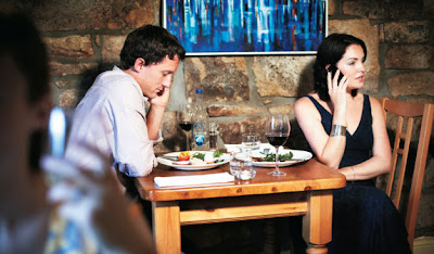 Foto de um casal a mesa de um restaurante, cada um falando em seu próprio celular