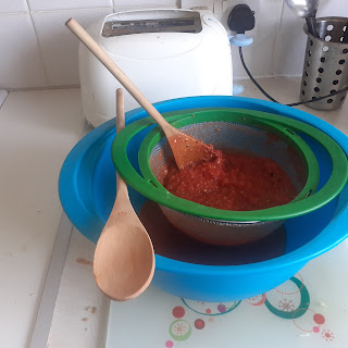 A large, plastic, blue bowl with a smaller green sieve balanced on top. The sieve is too small to balance on the bowl so one side is balanced on a wooden spoon across the bowl. Another wooden spoon is in the sieve to push the mixture through.