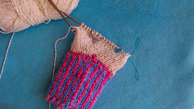 a white mitten-in-progress with a two-colour ribbed cuff in blue and pink.