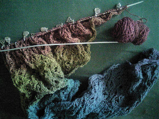 A lace shawl on a set of straight needles.  The yarn is a gradient from blue to red, and there is a small amount of yarn left in the cake of yarn.  There are a number of stitch markers with silver charms looped around the needles.