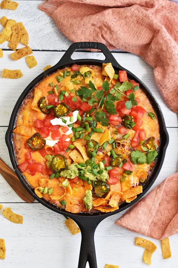 A delectable chili mixture topped with melted cheese, Fritos, fresh tomatoes, cowboy candy, green onions, sour cream, guacamole, and fresh cilantro in a cast iron skillet set on a white wood background