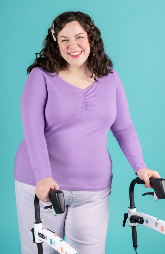 Model with brunette curly hair and holding mobility aid walker wears purple long sleeve jersey top with sweetheart neckline