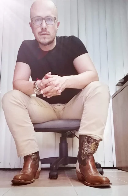 Bald dude wearing glasses with a black shirt on tucked into khaki pants wearing orange brown leather cowboy boots sitting about 3 ft from the camera center straight