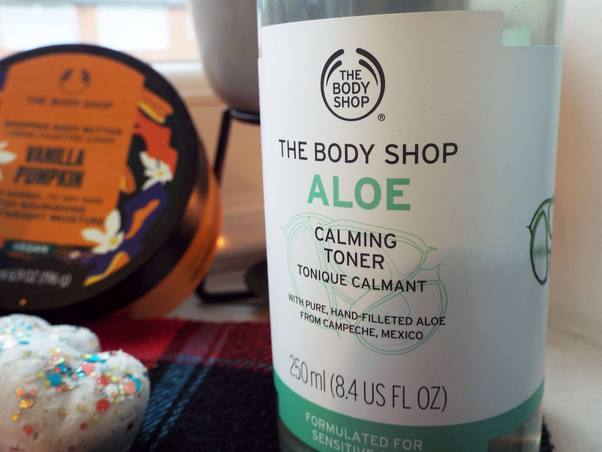 Close up of The Body Shop Aloe Toner, with Vanilla Pumpkin Body Butter and wax melts in background, on tartan scarf.