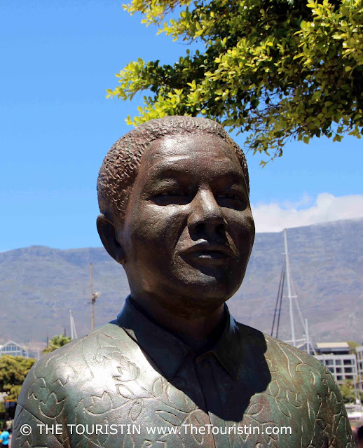 The upper half of a larger than life bronze statue of Nelson Mandela under a bright blue sky in front of a mountain.