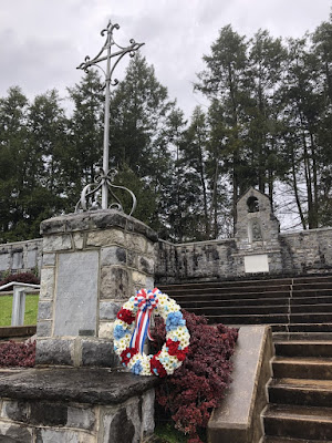 Lower left corner is stone pillar with a metal cross on top. A red, white, and blue wreath has been placed next to the pillar. Behind there are stone steps and a stone wall with plaques honoring 28th Division members who died in World War 1 and World War 2