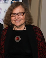 Mary Ellen Copeland at the Voice Awards. A smiling older woman with grey hair is wearing black-framed glasses, a black shirt, a red patterned kimono, and a round pendant with a red stone in the center. 