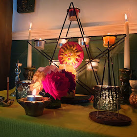 Green paneled wall, yellow altar cloth, pentagram candle-holder with sun plaque, home grown roses. Two green candle-holder jars. goddess shaped incense burner at the back. Two taper candles in green holders. The altar is a mostly symmetrical arrangement. Three incense sticks in a pentacle shaped flat incense burner. 