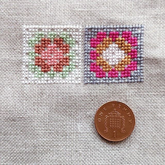 Two cross stitched granny squares with a penny on the fabric below. The granny squares are each just a bit larger than the penny