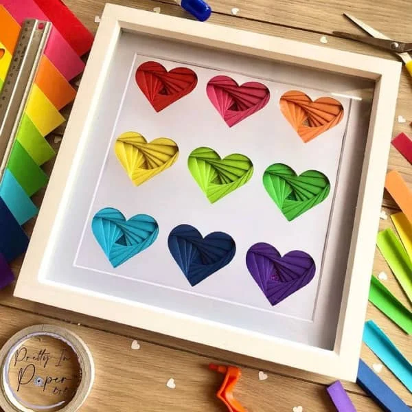 folded hearts in rainbow colors shown in square white frame surrounded by paper crafting supplies