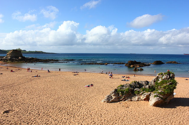 Letni wieczór na plaży El Camello w Santander. Piaszczysta plaża, na której znajduje się skała porośnięta roślinami. Przy linii brzegu wystają ponad taflę spokojnego morza mniejsze, czarne skały. Jedna z nich ma formę wielbłąda. Na plaży jest niewiele osób. 