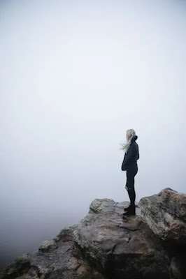 An image of a very alone sad girl standing at the mountain cliff in the fog.