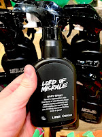 A white hand holding a black spray bottle filled with body spray with a jock arched label on the front that says Lord of Misrule body spray in white font on a background that’s bright and full of other black body spray bottles