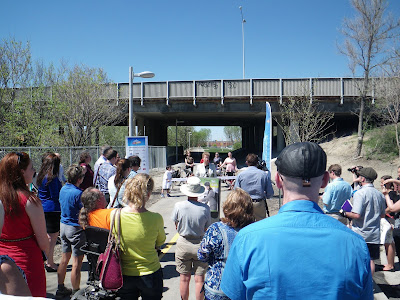 Looking along the O-Train pathway with the 417 overpass bridge across it, Diane Holmes standing at a podium speaking, with bicyclists standing behind her and a crowd in front.