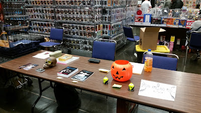 A vendor table at a comic book convention with two stacks of books, a fake skull and a Jack 'O Lantern candy bucket.