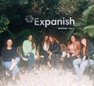 Seis mulheres estão sentadas em um jardim onde se lê uma placa escrito Expanish. São estudantes da escola de espanhol.