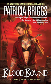 A woman with a bat tattooed on her chest and a paw on her stomach stands against a blood-red sky, holding a wrench. Bats are hanging above her and flying in the distance.