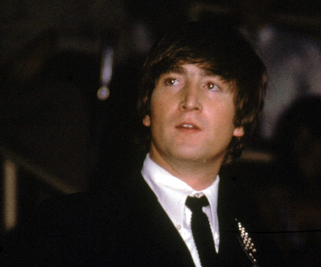 John Lennon viste un traje negro y toca una guitarra en un concierto de los Beatles a mediados de la década de 1960