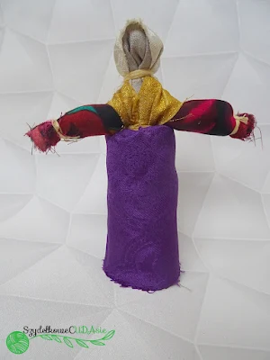 Lalka wykonana ze skrawków materiału ma lnianą głowę, beżową chustę na głowie, czerwone ręce, złotą bluzkę i fioletową spódnicę. Lalka taka nazywana jest motanką.