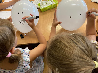 Na zdjeciu dzieci ozdabiające białe balony, czarnym pisakiem, dzieci mają odwrócone twarze, siedzą przy stole.