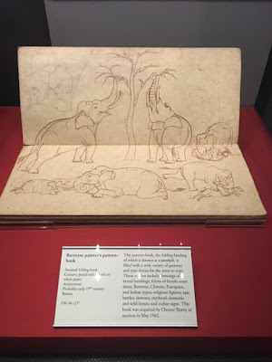 A narrow book, with the binding running horizontally and the upper page lifted at a right angle to the lower page and table, with fine umber ink sketches of elephants reaching up to a tree, laying doen, and calves playing.