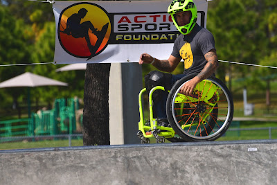 A manual wheelchair user is shown, gliding parallel to the lip of a ramp, waiting to drop in. He is wearing a bright green, motorcycle helmet, which matches the bright green frame of his wheelchair.