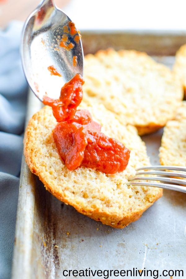 Spooning tomato based pizza sauce onto engish muffins - Use Canyon Bakehouse English muffins to make an easy crust for gluten free mini pizzas! They make a perfect pizza base for this easy kid friendly lunch recipe. Personalize them any way - even make them dairy free or vegetarian. Let kids choose their own healthy toppings for DIY personal pizza activity.