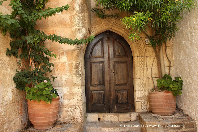 The dark wooden entrance door of a soft red-beige stone house framed by green plants.