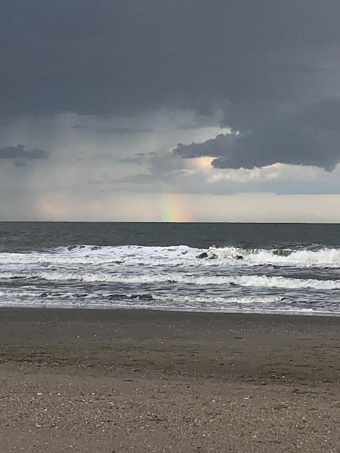 Looking out towards the endless ocean from the beach. White bubbles take up a third of the picture where the waves have crested and hit the beach. There are ominous, dark storm clouds hovering over the ocean. In the distance, towards the horizon is a section of rainbow barely seen, but there.