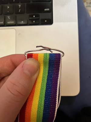 alt= The end of a pair of clip-on, rainbow elastic suspenders. The clip has been removed, leaving the wire that attached it to the elastic. The wire has been bent so that the ends that were holding the clip in place overlap each other. End ID.