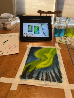 Ipad with a video playing, watercolor of the northern lights taped to the table, art supplies scattered around