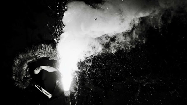 Black and white image of a Masked, hooded white man holding a blazing flare.