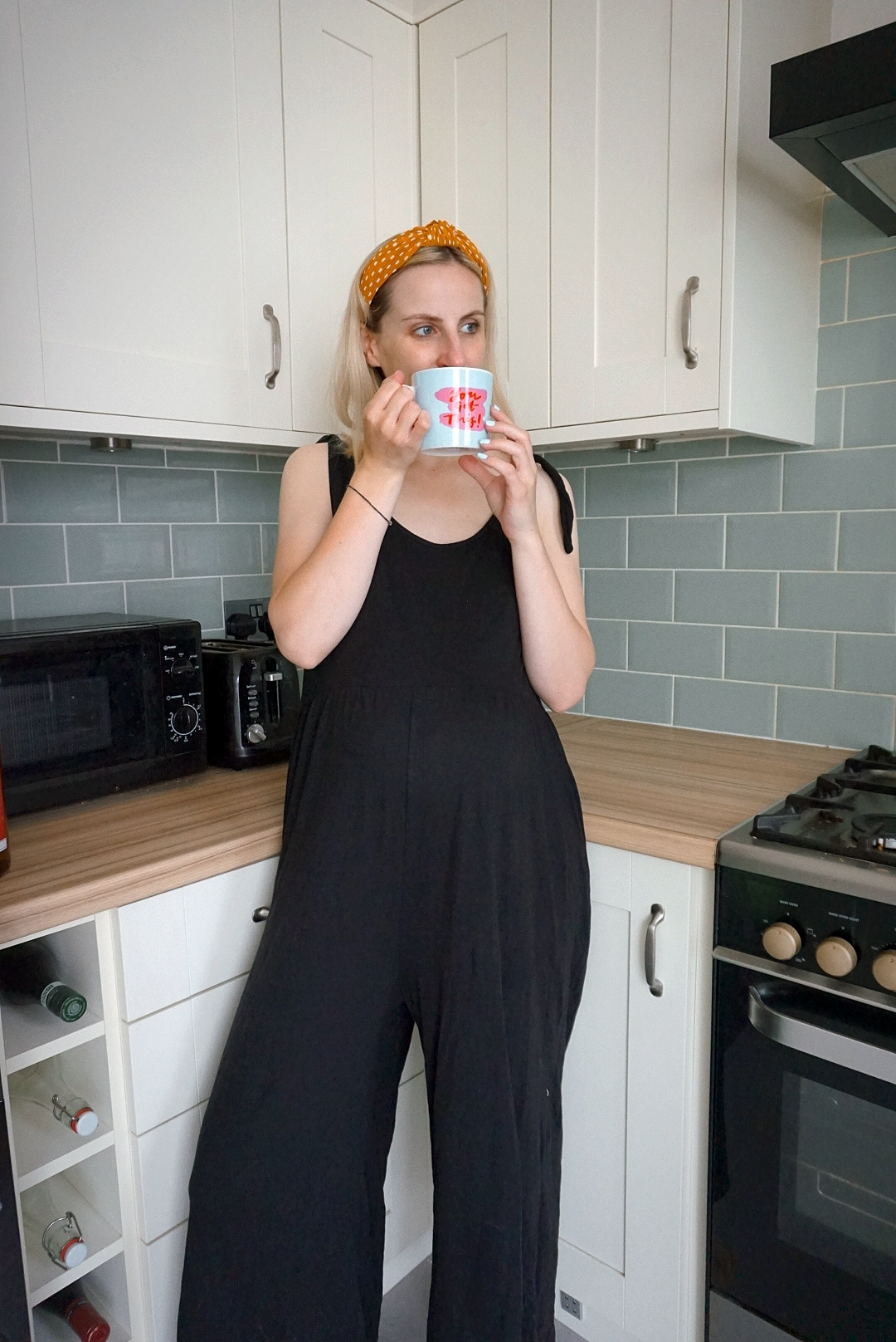 Outfit photo: Amy is stood in her kitchen with a mug wearing a black oversized sleeveless jumpsuit