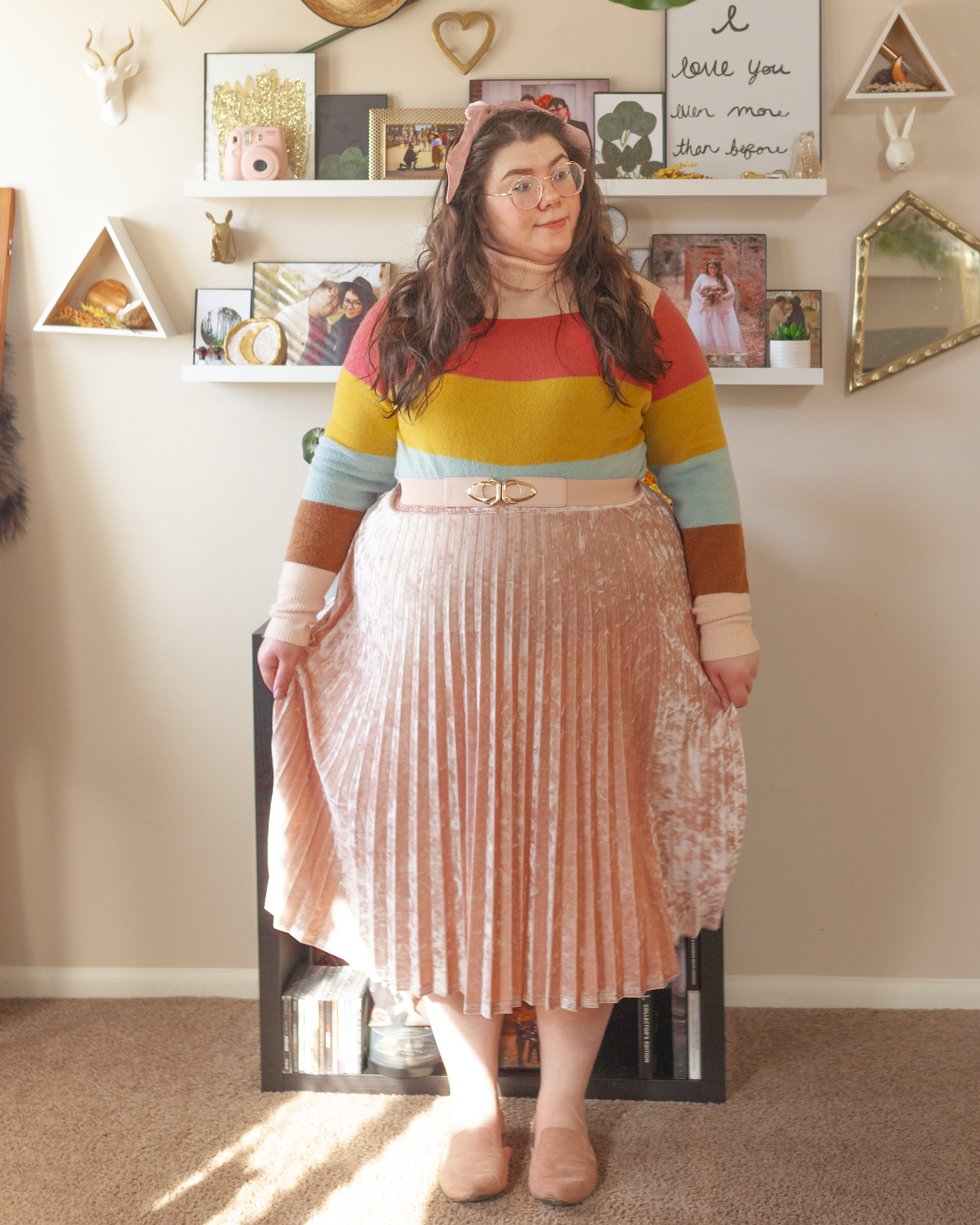 An outfit consisting of a light brown, pastel pink, pastel blue and marigold yellow turtleneck sweater tucked into a pastel pink crushed velvet pleated midi skirt.