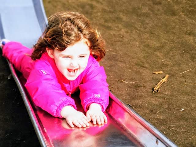 Image of a young girl laying on her front and sliding down a metal slide in an outdoor playground. The girl is wearing a bright pink Trespass all in one waterproof suit.
