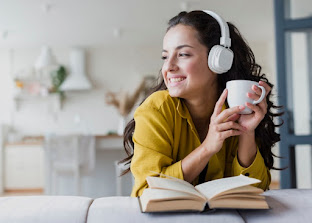 A caucasian woman with brown wavy hair wearing white headphones holding a cup with and open book in form of her