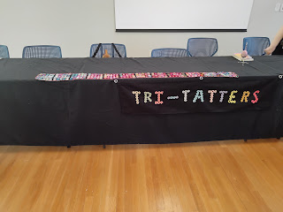 Table in open room with Tri-Tatters banner in front and scarf laid out on top, chairs are behind the table