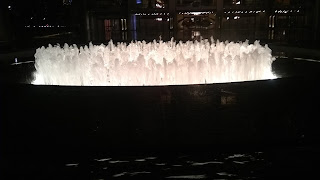 Photo taken at night. A circular fountain with upward gouts of water, in a circle,  lit up as if from within;