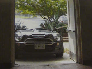 From inside a building, looking through an open two-door doorway. A black car sits just outside the threshold. A tree with green leaves can be seen in the background.