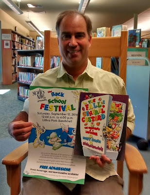 Ashland, Oregon author and educator Matt Damon in a rocking chair in Bellview library, holding a poster to promote the Back to School Festival and a copy of his book, 'The Fall of General Custard: Or the Overthrow of a Leftover'