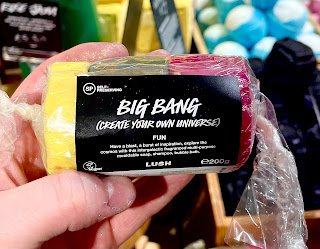 A photo of a white hand holding a long cylindrical log of blue red and yellow fun wrapped in clear packaging with a black rectangular label that says big bang (create your own universe) fun lush in white font on a bright background