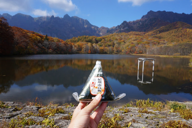 An onigiri held in front of the Kagami Ike (Kagami Lake) that features orange fall leaves in Togakushi, Japan