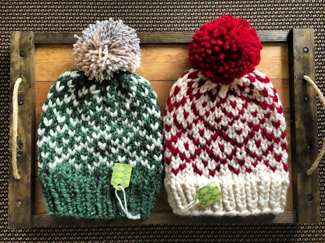 Two matching knit hats next to each other. Left one is green with cream accents and a neutral pom. Right one is cream with red accents and a red pom. They're flat laying on a wooden tray. Each have gift tags attached to them.