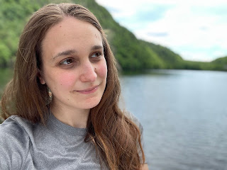 A person with long, brown hair is wearing a grey shirt and standing in front of a lake