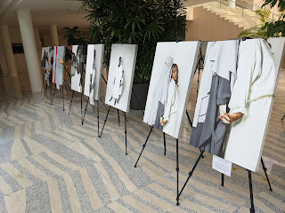 Photo by Rob Goetze of part of the Anti-Islamophobia exhibit. Seven large photos, each on a stand, in the large hall at Edmonton City Hall. Two photos that are closer in view are of a Middle Eastern woman wearing a burka standing back to back with a Black woman in a white dress, hands clasped together, and another of a Black woman with her daughter peering out from behind her.